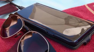 Güneş gözlüklerindeki bulanık yansımalar ve kumsaldaki kırmızı havlunun üstündeki akıllı telefon ekranı.
