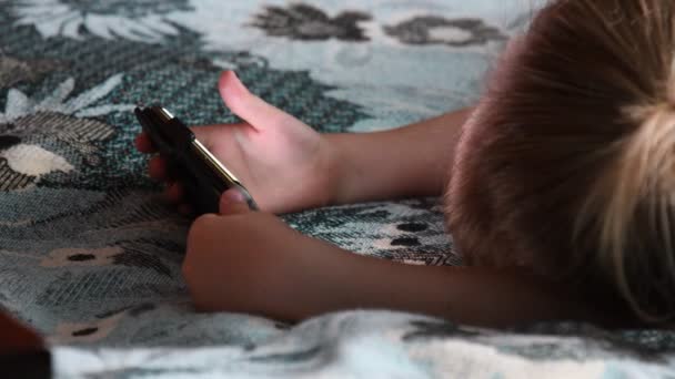 Enfant regardant à l'écran du smartphone tenant le dispositif dans les mains et couché à la couverture — Video