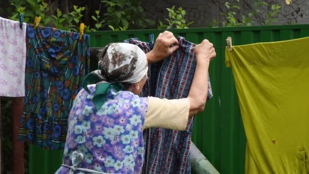 Kırsal kesimdeki yaşlı bir kadın çamaşırları dışarıdaki mandallara asıyor. — Stok video