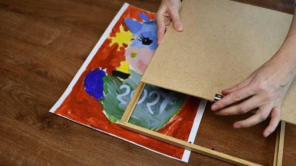 Руки женщины помещают детскую живопись в деревянную рамку с фанерным основанием — стоковое фото
