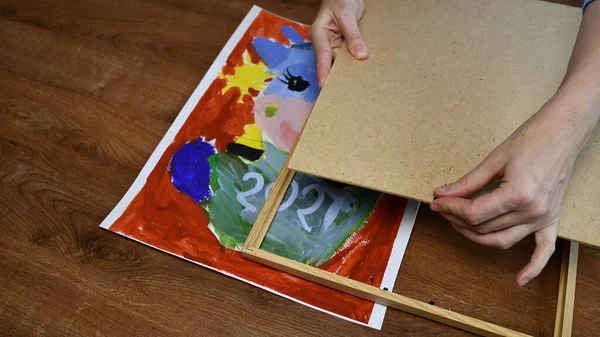 Руки женщины помещают детскую живопись в деревянную рамку с фанерным основанием — стоковое фото