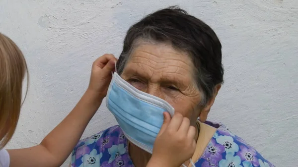 Toddler gir handsl helping her great-grandmother to put on disposable medical mask to wrinkled face — Stock Photo, Image