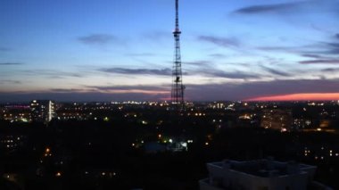 Lacivert gökyüzü, dönen gri bulutlar ve yanıp sönen şehir ışıklarıyla iletişim kulesinin silüeti.