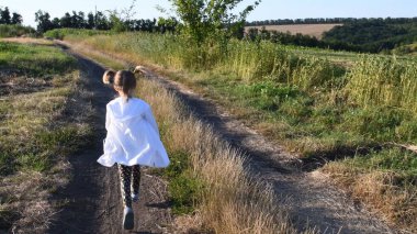 Küçük kız yaz manzarasının arasında köy yolunda koşuyor.