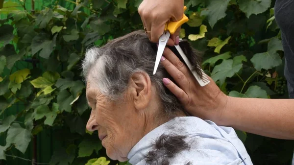 Las manos femeninas con peine y tijeras cortan el cabello de la persona mayor —  Fotos de Stock