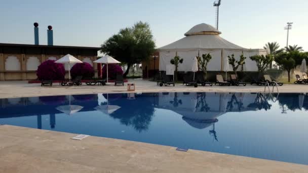 Água da piscina calma no resort de luxo com espreguiçadeiras vazias e guarda-sóis brancos à beira da piscina — Vídeo de Stock