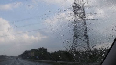 Arabanın ön camında yağmur damlaları ve bulanık şanzıman kulesi silueti var.