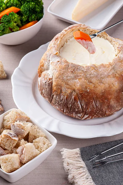 Creamy cheese fondue inside Italian bread. Fork dipping carrot into cream cheese