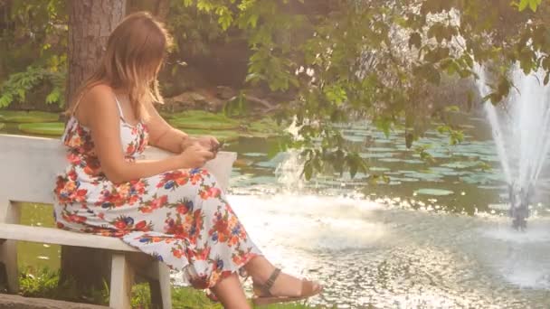 Девушка на скамейке возле фонтана проверяет телефон — стоковое видео