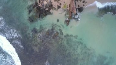 Köpüren okyanus dalgaları gri kayalıklarla kumlu sahil şeridinde yuvarlanıyor.