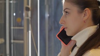Genç bir kadın, hırdavatçıdaki duşta akıllı telefondan konuşur.