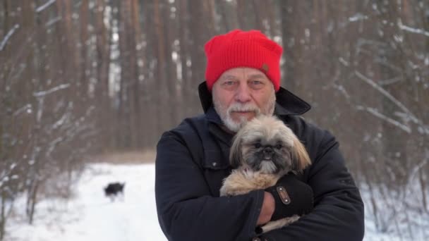 Leeftijd man met rode hoed houdt hond in de armen en loopt langs het bos — Stockvideo