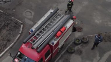 İtfaiyeciler aracın ve paslı kanalizasyon kapağının yanında dururlar.