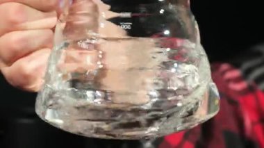 İnsan eli sıvı içerikli şeffaf bir kap tutar. Dalgalı yüzeyi olan cam objeyi kavrayan kişi yavaş çekimde bozulmalar yaratır.