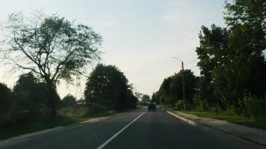 Ön cam manzarası uzun ağaçlar arasında düz asfalt yolu gösteriyor. Akşam güneşi parlarken küçük araba beyaz merkezli yol gösterici şeridi ile ilerliyor.