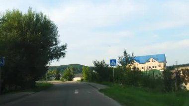 Ön cam manzarası dar şeritli sessiz köy köşesini gösteriyor. Beyaz çitler ve mavi çatı evi kayarken yol kenarındaki ağaçlar yumuşak gölgeler bırakıyor.