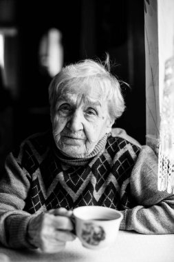 Yaşlı bir kadın evde kahve içer. Siyah beyaz fotoğraf.