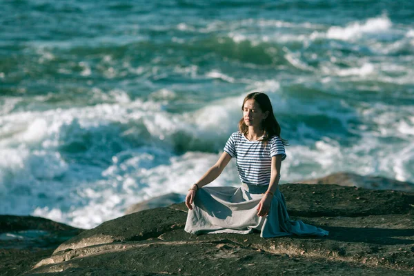 若いですヨガ女性Medatesで蓮の位置に座って岩の上に海の海岸 — ストック写真