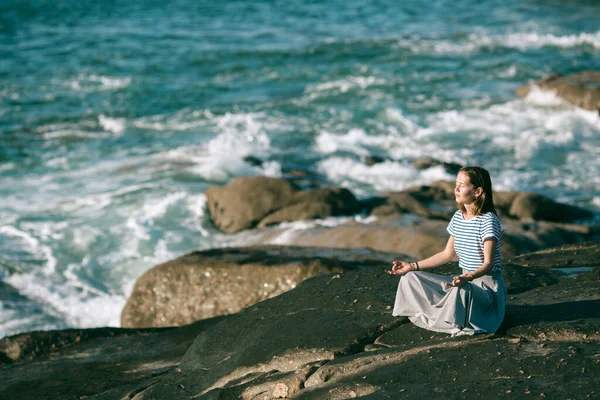 若いですヨガ女性Medatesで蓮の位置に座って岩の上に大西洋の海岸 — ストック写真