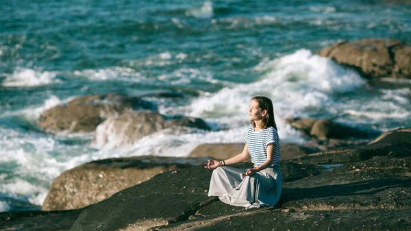 若いですヨガ女性Medatesで蓮の位置に座って岩の上に大西洋海岸 — ストック写真