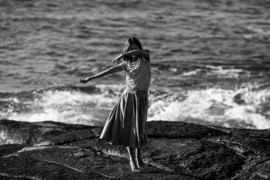 Genç dansçı kadın okyanus kıyısında koreografiyle meşgul. Siyah beyaz fotoğraf.