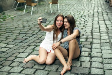 İki kız arkadaş, eski kasabadaki kaldırımda oturmuş akıllı bir telefondan selfie çekiyor.