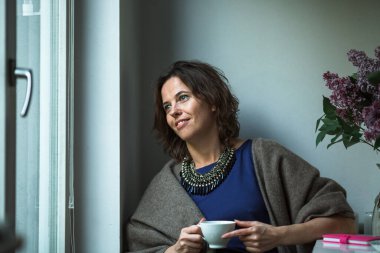 Pencereden dışarı bakıp elinde bir fincan çay tutan bir kadın..