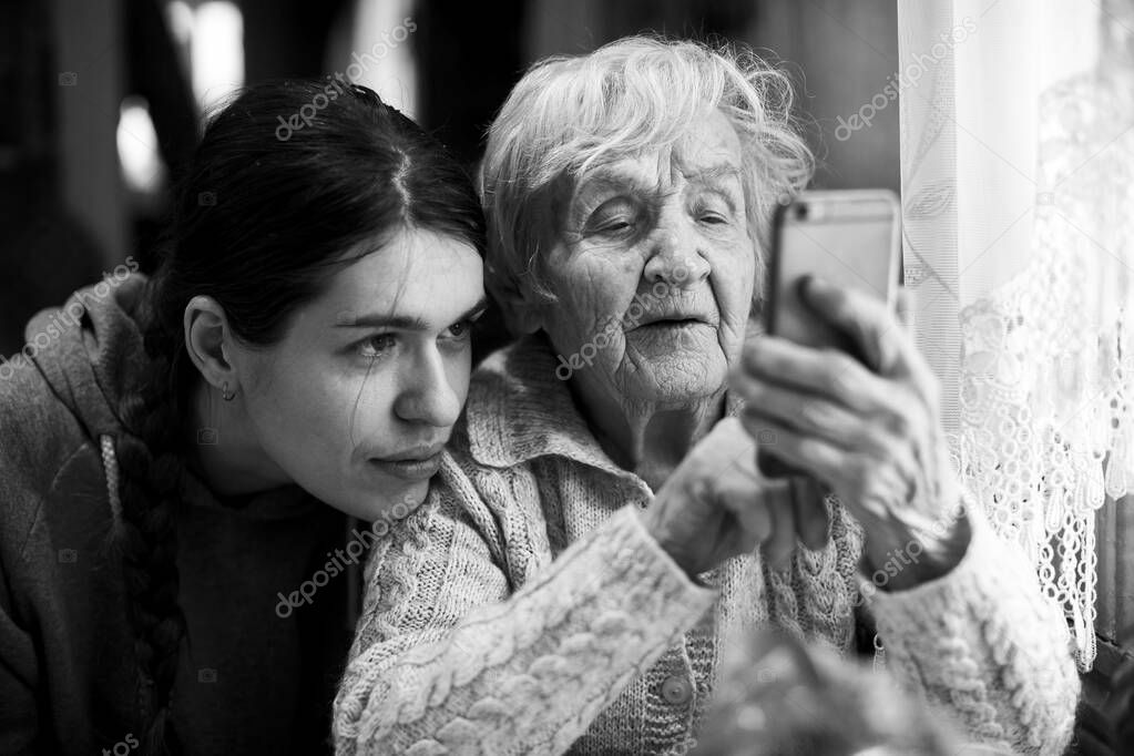 An old woman looks at a smartphone, with his adult granddaughter. Black and white photo.