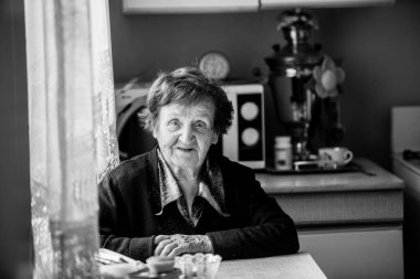 Yaşlı bir kadın mutfaktaki evinde oturuyor. Siyah beyaz fotoğraf.