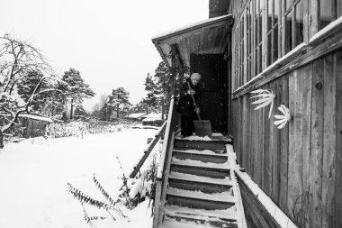 Yaşlı bir kadın kırsal evinin verandasındaki karı temizliyor. Siyah beyaz fotoğraf.