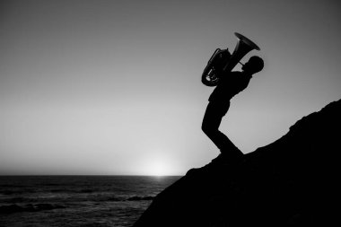 Deniz kıyısında tubası olan bir müzisyenin silueti. Siyah beyaz fotoğraf.