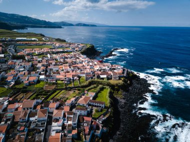 Atlantik Okyanusu 'nun üzerindeki yeşil kayalıklarda kırmızı çatı evleri olan Azores' de bir sahil kasabası. Dalgalar parlak bir gökyüzünün altındaki kayalık sahile vurur, havadan görülür..