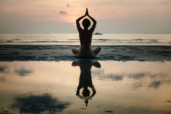 Силуэт женщины, медитирующей в позе йоги на пляже на рассвете, руки подняты над головой, отражены в мелководье, спокойном море и мягком утреннем свете.