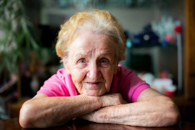 Yaşlı bir kadının kollarını masaya dayayıp nazikçe gülümseyerek kameraya bakışının yakın plan portresi. Kapalı alan ışığı yüzünü ve kırışıklıklarını aydınlatıyor..