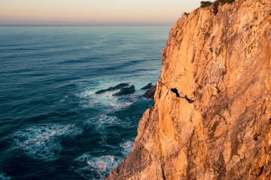 Derin mavi Atlantik sularının üzerindeki engebeli okyanus uçurumunda altın saat ışığı. Dalgalar karanlık kıyı kayalarına çarpıyor..
