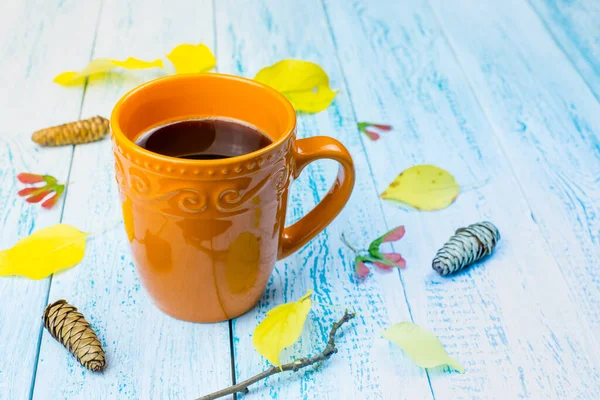 A cup of coffee with autumn leaves. Autumn background. Coffee on a blue board background with yellow falling leaves and cones. Hello, autumn.