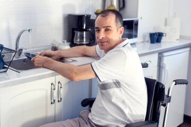 Young man Disabled Man On Wheelchair Washing Dishes. Smiling Young Handicapped Man Sitting On Wheelchair In Kitchen. Focus on his face.