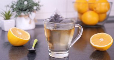 Yakınlarda limonları olan, mutfak tezgahındaki bir fincanın içindeki sıcak suya batırılan ve kaldırılan çay poşetinin yakın çekimi. Çay demleme süreci, rahat ve rahatlatıcı an.