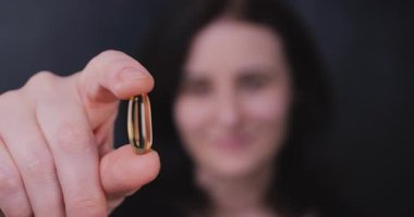 Yetişkin bir kadın kameraya doğru omega-3 hapı tutuyor, kalp sağlığı, takviyeler, kendine bakım ve genel olarak sağlığın altını çiziyor..