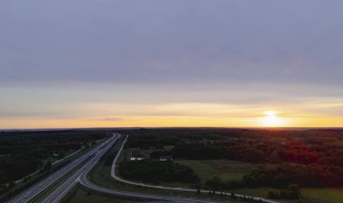 Renkli bir gün batımının altında sarmal bir otoyolun hava manzarası yemyeşil tarlalarla çevrili. Doğanın güzelliğini ve gün batımında ulaşımı gösteriyor..