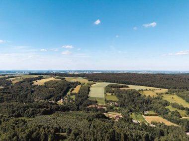 Açık mavi gökyüzünün altındaki geniş yeşil tarlaların ve ormanların havadan görünüşü doğa ile çevrili tarım arazilerini ve kırsal evleri gösteriyor..