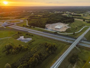 Gün batımında bir otoyol kavşağının havadan görünüşü yemyeşil ve şehir altyapısının genişlemesini gösteren bir inşaat alanı ile çevrilidir..