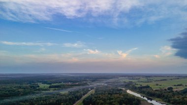 Bereketli yeşilliklerin ve yumuşak bulutlarla dolu berrak bir gökyüzünün altında dolambaçlı bir nehrin yer aldığı uçsuz bucaksız bir manzara. Doğa ve seyahat temaları için ideal.