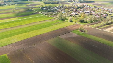 İlkbaharda yakınlardaki bir köy tarafından tamamlanan canlı yeşil tarlaları ve kahverengi toprak örneklerini gösteren kırsal bir manzaranın havadan görünüşü tarımsal güzelliği vurguluyor..