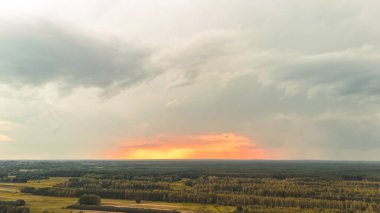 Canlı bir günbatımının havadan görünüşü yeşil tarlalar ve ormanlık alanlar üzerindeki ufku aydınlatıyor. Dramatik bulutlar renkli gökyüzüne derinlik katar.