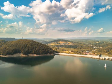 Polonya 'daki Solina Barajı' nın hava manzarası yemyeşil ve durgun sularla çevrili. Mavi gökyüzü altında beyaz bulutlar var. Doğa ve seyahat temaları için ideal.