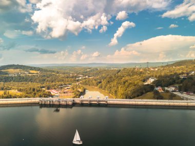 Solina Barajı Polonya 'nın yemyeşil yeşilliklerle çevrili nefes kesici hava manzarası ve dingin bir göl, canlı bir gökyüzü altında sakin sularda yüzen bir yelkenliyi gösteriyor..