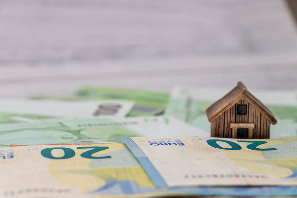 A miniature wooden house sits atop a stack of 20 Euro banknotes representing concepts of mortgage finance real estate investment and homeownership in a financial setting.