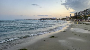 Alicante İspanya - 12 Temmuz 2025: Alicante plajında gün batımı manzarası hafif dalgalar kıyıda renkli gökyüzüne ve kıyı şeridi boyunca hareketli atmosfere çarpıyor.