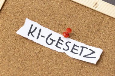 El yazısıyla yazılmış bir notta mantar panoya tutturulmuş KI-GESETZ yazıyor. Bu kavram, teknolojideki yeniliği sembolize eden yapay zeka düzenlemeleri ile ilgilidir..